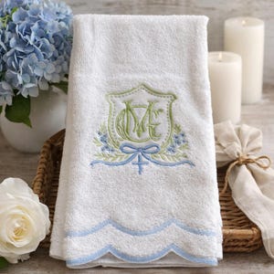 May include: White hand towel with a scalloped light blue border. The towel features an embroidered crest with the letters "ME" in green and blue, surrounded by floral and bow details. The towel is made of a soft, absorbent material.