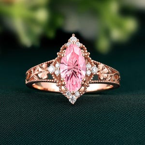 Könnte beinhalten: Ein roségoldener Ring mit einem rosa Stein im Marquise-Schliff, umgeben von kleinen weißen Edelsteinen. Das Band hat ein Blumenmuster. Der Ring ist vor einem dunkelgrünen Hintergrund platziert.