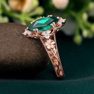 Può includere: Anello in oro rosa con una grande gemma verde taglio smeraldo. L'anello è impreziosito da piccole pietre chiare e dettagli floreali sulla fascia. L'incastonatura presenta un design decorativo in stile filigrana.