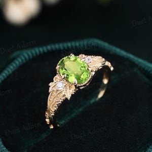 Könnte beinhalten: Ein goldener Ring mit einem zentralen, oval geschliffenen, hellgrünen Peridot, der von vier Krappen gehalten wird. Die Ringschiene ist mit detaillierten Blatt- und Rankengravuren verziert und hat zwei kleine, runde, klare Akzentsteine an den Seiten.