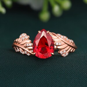 May include: A rose gold ring with a striking, pear-cut, deep red gemstone. The ring has feather-shaped details, embellished with small, sparkling white stones. The design is elegant and inspired by nature.
