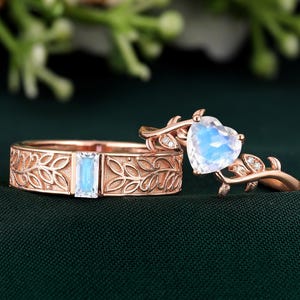 May include: Two rose gold rings. One ring features a rectangular, light blue gemstone set in a band with a floral design. The other ring has a heart-shaped, light blue gemstone, with small clear stones and a leaf design.