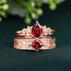 May include: A pair of rose gold rings with intricate leaf designs. The top ring features a pear-shaped garnet gemstone surrounded by smaller diamonds. The bottom ring has a square-shaped garnet gemstone.