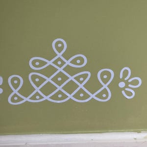 Op de afbeelding: Wit geometrisch ontwerp geschilderd op een lichtgroene muur. Het ontwerp heeft een centraal ruitvormig patroon met stippen en gebogen lijnen, geflankeerd door bloemmotieven. De muur heeft een zachte, gedempte groene kleur.