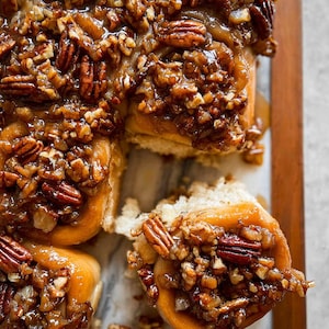 Könnte beinhalten: Draufsicht auf Pekannuss-Karamell-Brötchen. Die goldbraunen Brötchen sind mit gehackten Pekannüssen und einer glänzenden Karamellglasur bedeckt. Einige Brötchen sind angeschnitten und zeigen das weiche Innere.