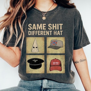 May include: Dark grey t-shirt featuring the text "SAME SHIT DIFFERENT HAT" above four images of different hats. The hats include a white hood, a grey cap with a Confederate flag, a black military hat, and a red "Make America Great Again" hat.