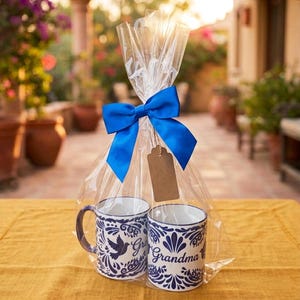 May include: Two white ceramic mugs with blue floral and bird designs, wrapped in clear cellophane and tied with a blue ribbon. One mug says "Grandma". A blank tag hangs from the ribbon.