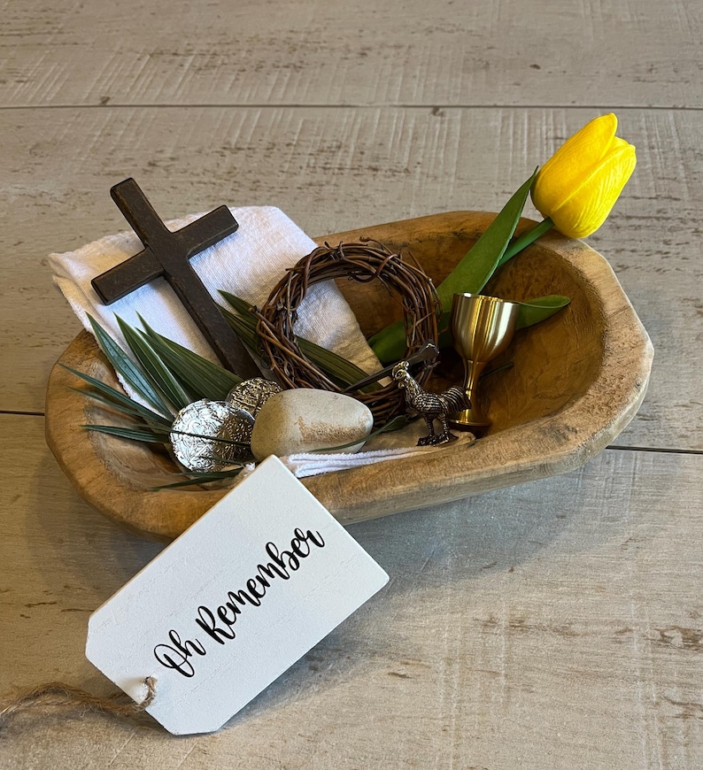 May include: A wooden bowl filled with religious symbols, including a cross, a crown of thorns, a chalice, a rooster figurine, and a palm branch. A white tag with the text "Oh Remember" is attached to the bowl. A yellow tulip is in the background.
