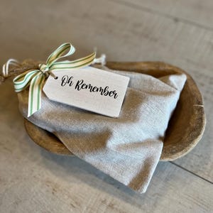 May include: A wooden bowl with a white fabric bag inside. The bag is tied with a green and white striped ribbon and a white tag that says "Oh Remember".