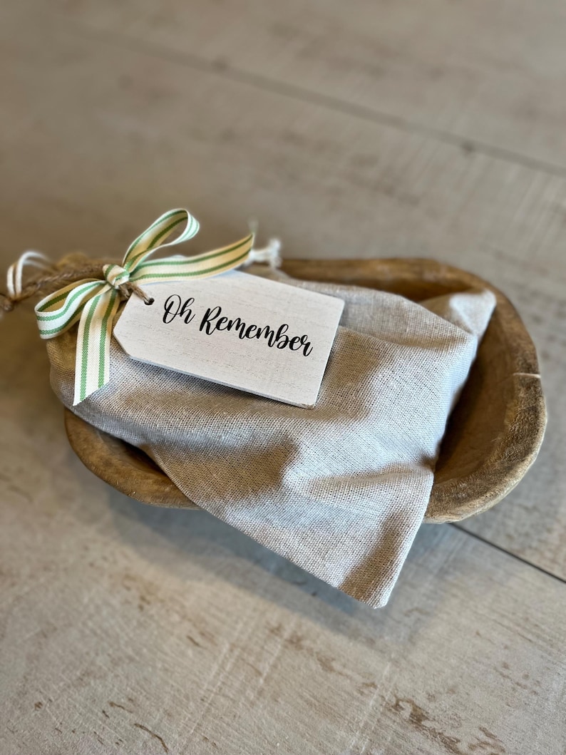 May include: A wooden bowl with a white fabric bag inside. The bag is tied with a green and white striped ribbon and a white tag that says "Oh Remember".