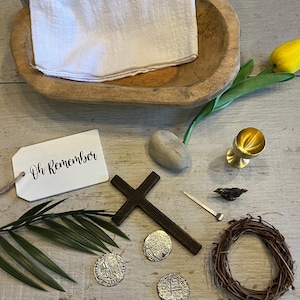 May include: A wooden bowl with a white cloth inside, a cross, a crown of thorns, three silver coins, a nail, a small bird, a stone, a gold chalice, a palm branch, and a white tag with the text "Oh Remember" on it.