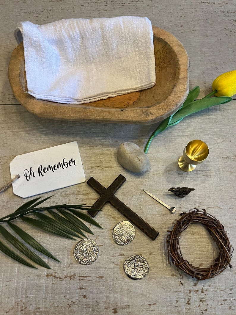 May include: A wooden bowl with a white cloth inside, a cross, a crown of thorns, three silver coins, a nail, a small bird, a stone, a gold chalice, a palm branch, and a white tag with the text "Oh Remember" on it.