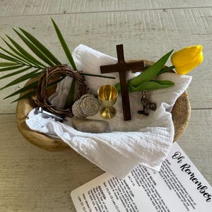 May include: A wooden bowl with a white cloth inside. The bowl contains a variety of objects, including a small wooden cross, a golden chalice, a rooster figurine, a wreath of twigs, a stone, and a yellow tulip. A card with the text "Oh Remember" is visible in the foreground.
