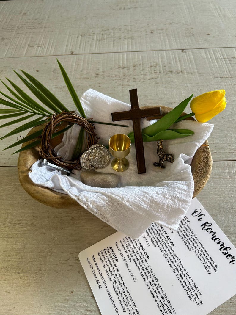 May include: A wooden bowl with a white cloth inside. The bowl contains a variety of objects, including a small wooden cross, a golden chalice, a rooster figurine, a wreath of twigs, a stone, and a yellow tulip. A card with the text "Oh Remember" is visible in the foreground.