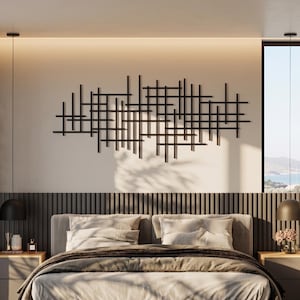Op de afbeelding: Een groot zwart metalen wandobject bestaande uit een raster van elkaar kruisende horizontale en verticale lijnen. Het kunstwerk is bevestigd aan een lichte muur boven een bed en vormt een geometrisch patroon in het midden van de muur.