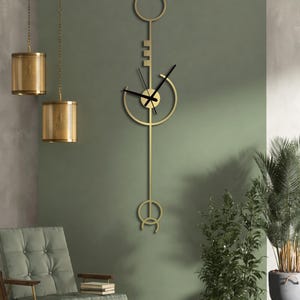 May include: A gold-coloured, key-shaped wall clock with black clock hands hangs on a green wall. The clock has a circular face and a long, vertical stem with decorative elements. The room also features a chair, plants, and hanging gold lamps.