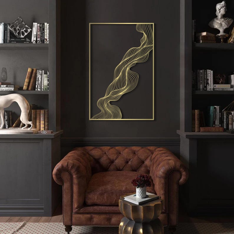 May include: A gold-framed wall art piece with a dark background. The artwork features a flowing, abstract design in gold lines. The piece is displayed above a brown leather armchair in a room with dark gray walls and bookshelves.