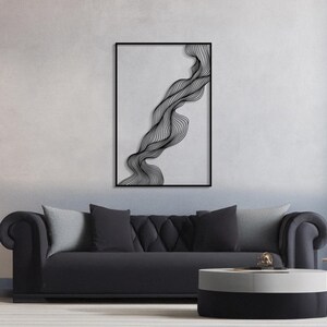 May include: Black framed wall art featuring a flowing, abstract design of black lines on a light gray background. The artwork is rectangular and hangs above a black sofa with gray pillows. A round, two-tone coffee table sits in front of the sofa.