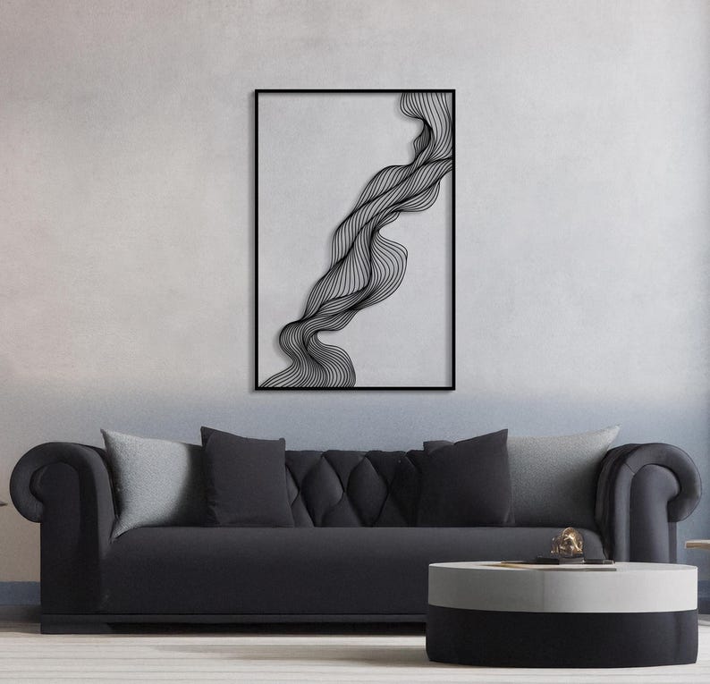 May include: Black framed wall art featuring a flowing, abstract design of black lines on a light gray background. The artwork is rectangular and hangs above a black sofa with gray pillows. A round, two-tone coffee table sits in front of the sofa.