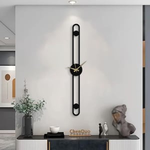 Puede incluir: Un reloj de pared moderno negro con agujas doradas y una cara circular. El reloj está montado en una pared blanca con un marco negro alrededor. El reloj está en un salón con una mesa y un jarrón de flores.