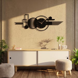 Op de afbeelding: Een wandklok van zwart metaal met een horizontale opstelling van geometrische vormen, waaronder cirkels, rechthoeken en parallelle lijnen. De centrale ronde wijzerplaat toont Romeinse cijfers en dunne goudkleurige wijzers.