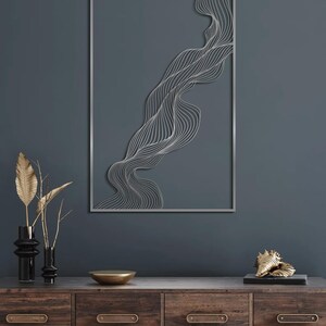 May include: A silver metal wall art piece with a flowing, abstract design is mounted on a dark teal wall. The artwork is rectangular with a thin silver frame. A wooden cabinet with decorative items, including vases and a gold bowl, sits below the artwork.