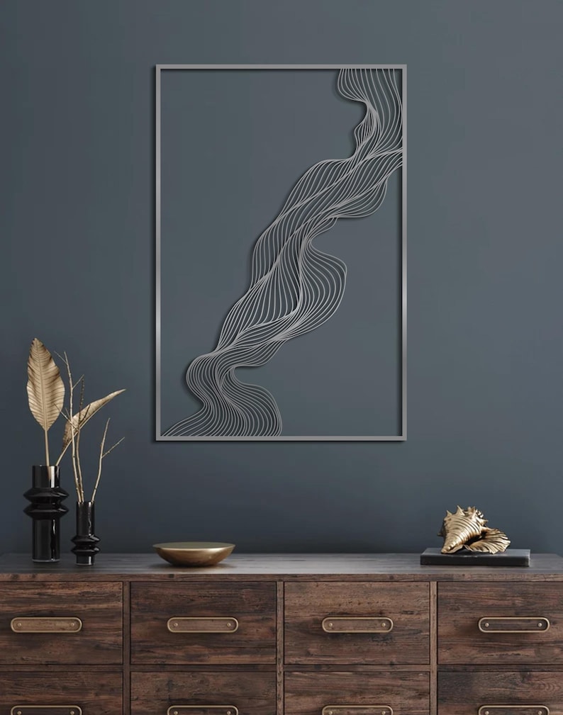 May include: A silver metal wall art piece with a flowing, abstract design is mounted on a dark teal wall. The artwork is rectangular with a thin silver frame. A wooden cabinet with decorative items, including vases and a gold bowl, sits below the artwork.