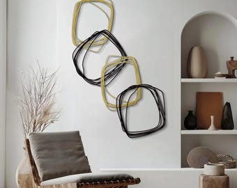 May include: Abstract wall art featuring four overlapping, irregular square shapes. Two shapes are outlined in black, and two are outlined in gold. The art is mounted on a white wall, creating a modern, minimalist aesthetic. A wooden chair and decorative items are in the foreground.