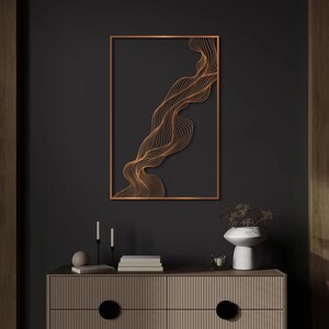May include: A copper-colored, abstract wall art piece featuring a flowing, wave-like design within a rectangular frame. The artwork is mounted on a dark gray wall, with a wooden cabinet and decorative items below.