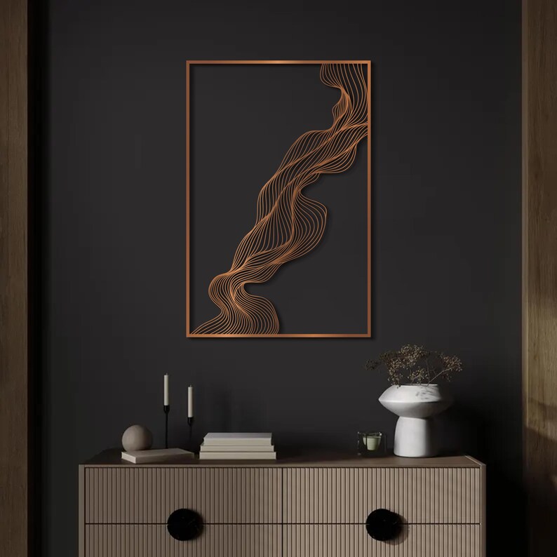 May include: A copper-colored, abstract wall art piece featuring a flowing, wave-like design within a rectangular frame. The artwork is mounted on a dark gray wall, with a wooden cabinet and decorative items below.
