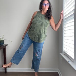 May include: An olive green sleeveless tank top featuring a dark green botanical leaf print pattern paired with blue denim jeans that have colorful painted floral accents on the legs and brown flip-flop sandals on the feet.