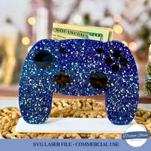 May include: A blue and silver glitter-covered controller-shaped money holder. The controller has black buttons and a cross-shaped directional pad. A twenty-dollar bill is visible in the slot. The text "SVG LASER FILE + COMMERCIAL USE" is at the bottom.
