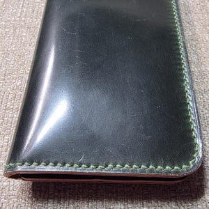 May include: A rectangular black leather wallet featuring a smooth, polished surface and contrasting lime green stitching along the edges. The wallet has a slim, folded design with visible layers of leather along the bottom edge.