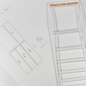 May include: A clear plastic fraction slices tool next to a sheet of white paper with handwritten math problems. The paper shows a fraction addition problem: 1/2 + 1/4 = 3/4. The tool is labeled "FRACTION SLICES".