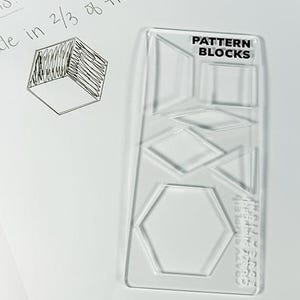 May include: A clear plastic Pattern Blocks template with various geometric shapes, including a hexagon, diamonds, and a cube. The template is on a white surface with handwritten text that reads "Fractions! 1. Shade in 2/3 of the Hexagon below."