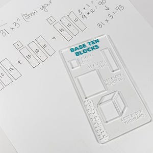 May include: A clear plastic Base Ten Blocks tool rests on a white sheet of paper with handwritten math problems. The text reads "Multiplication! What is 31 x 3?" with the answer shown. The tool has the words "Base Ten Blocks" printed on it.