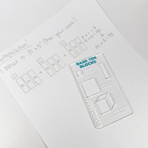 May include: A clear plastic Base Ten Blocks tool rests on a white sheet of paper with handwritten math problems. The text reads "Multiplication! What is 31 x 3?" with the answer shown. The tool has the words "Base Ten Blocks" printed on it.