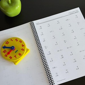 May include: A spiral-bound math fluency quiz for grade 4 with printed arithmetic problems, a yellow plastic analog clock with blue and red hands, and a glossy green ceramic apple figurine. The quiz page includes the text MATH FLUENCY QUIZ GRADE 4.
