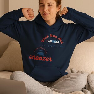 Puede incluir: Sudadera con capucha azul marino con bolsillo delantero y capucha con cordón. El gráfico en la parte delantera dice "Work From Bed, Inc." con una figura durmiendo y la palabra "snoozer" en rojo. La persona está sentada en una cama con un portátil y una taza.