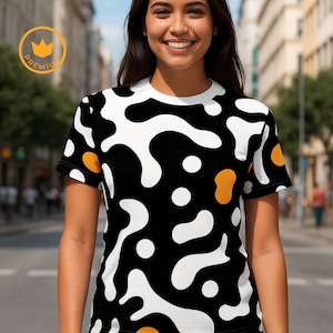 May include: A black t-shirt with a white and orange abstract pattern. The shirt has a crew neck and short sleeves. The design features organic shapes and dots. The shirt is being worn by a person in an outdoor setting.