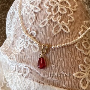 May include: A delicate pearl necklace with a gold clasp and a red gemstone pendant. The necklace is set against a backdrop of white lace with floral patterns. The word "EDELINE" is visible in the bottom right corner.
