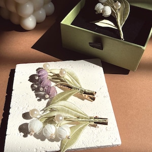 May include: Hair clips and a brooch featuring lily of the valley designs. The clips have white and purple flower buds with pearl accents and gold-tone clasps. A green box contains a brooch with similar floral details. The items are on a textured white surface.