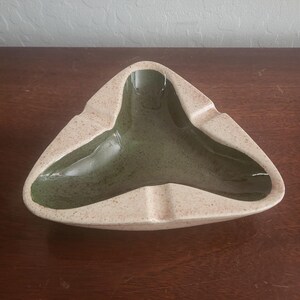 May include: A triangular ceramic ashtray featuring a smooth, dark olive green interior basin and a speckled, light beige exterior rim. The design includes three distinct notches along the edges, creating a symmetrical, three-pointed shape.