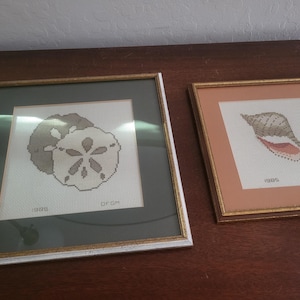 May include: Two framed needlepoint artworks featuring sea life. One depicts a sand dollar, the other a seashell. Both are framed in gold and silver-toned frames. The artwork includes the year "1985" and the initials "DFGH".