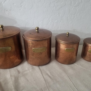 May include: A set of four graduated cylindrical copper canisters with matching lids, each topped with a small brass knob. Every canister features a rectangular brass label plate on the front, displaying engraved text for kitchen storage organization.