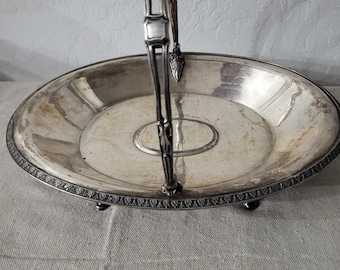 May include: A silver-toned serving tray with a decorative handle. The tray has a round base with an ornate edge and three small feet. The handle is attached to the center of the tray and features a decorative design at the top.