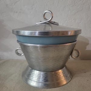 May include: A vintage, two-piece serving dish with a silver-colored metal exterior and a light blue interior. The lid has a loop handle. The dish has a hammered texture and two small handles on the sides.