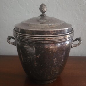 May include: A silver-colored, lidded ice bucket with two side handles. The lid is topped with a decorative finial. The bucket has a slightly distressed appearance, suggesting age or use. The bucket is sitting on a wooden surface.