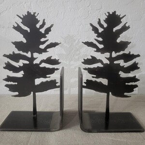 May include: Two black metal bookends, each featuring a silhouette of a pine tree. The tree design is detailed, with branches and needles. The bookends have a rectangular base and a vertical support.