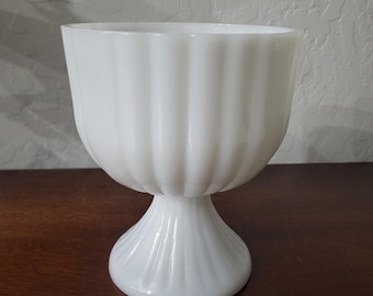 May include: White milk glass pedestal bowl with a fluted design. The bowl is on a wooden surface.